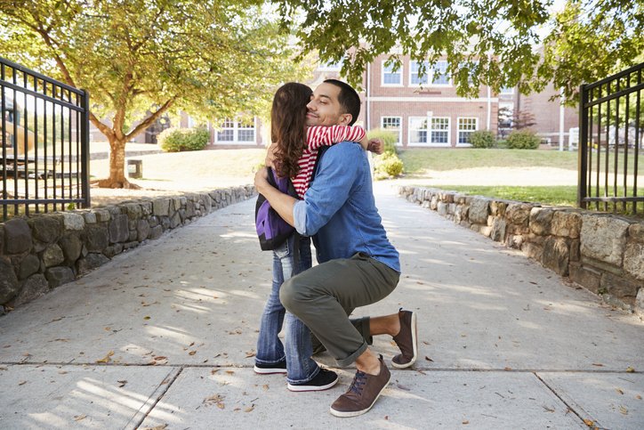 Parent kneeling to hug a child goodbye outside a school entrance