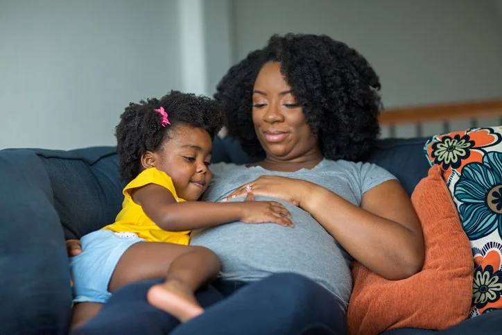 Toddler hugging a pregnant parent's belly while sitting together on the couch