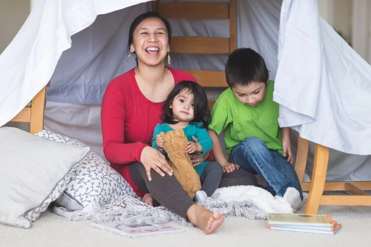 Parent and two children laughing together inside an indoor blanket fort