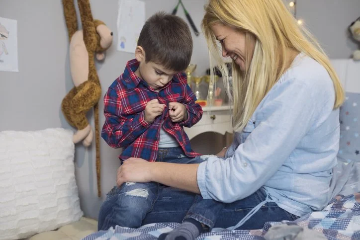 Parent helping a child button their shirt in the bedroom