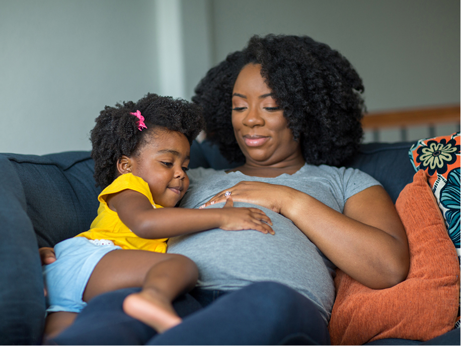 Toddler gently touching a pregnant parent's belly while sitting together on the couch