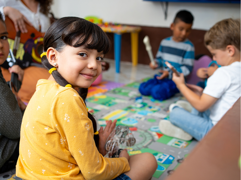 Child smiling in a preschool classroom while classmates play nearby